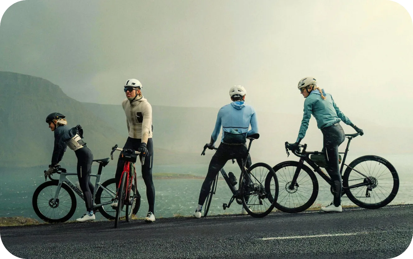 Four cyclists in gear pause with their bikes beside a misty coastal road overlooking water and cliffs.