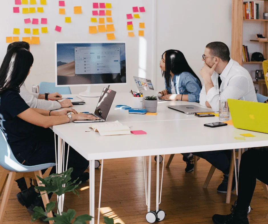 Team of four colleagues collaborating at a table with laptops, a monitor, and colorful sticky notes on the wall.