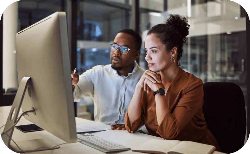 Two colleagues reviewing data on a desktop monitor, collaborating on AI reporting insights in an office.