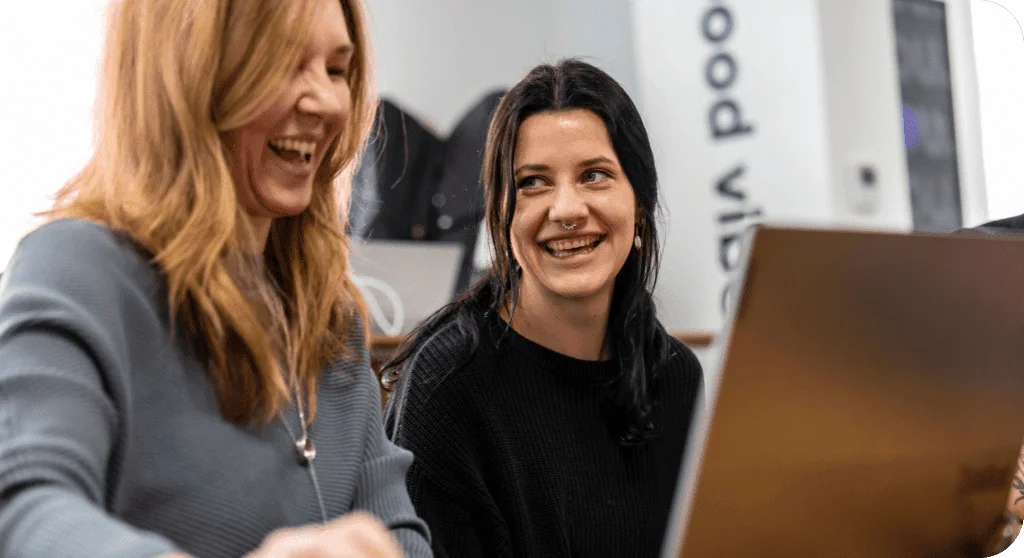 Two women laughing together while looking at a laptop screen in a bright office setting.
