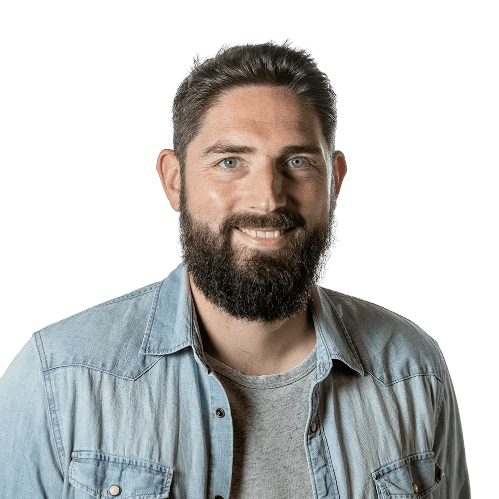 Smiling bearded man with short dark hair, wearing a light denim shirt, posed for a studio headshot.