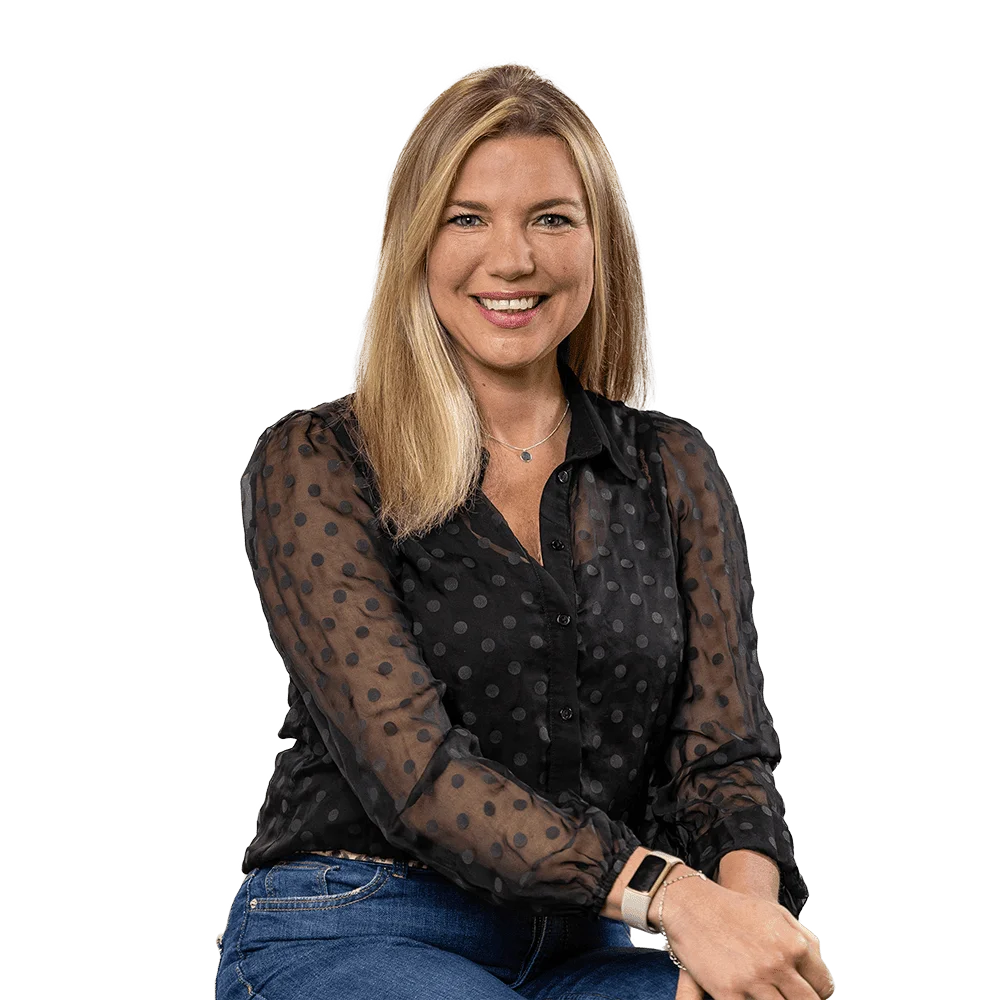 Smiling woman with shoulder-length blonde hair wearing a sheer black polka-dot blouse and jeans, seated against a white background.
