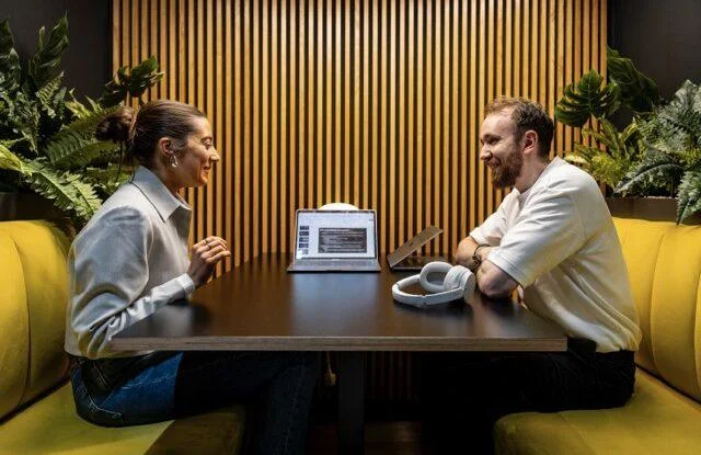 Two coworkers smiling in conversation at a table with a laptop and headphones in a modern office booth.