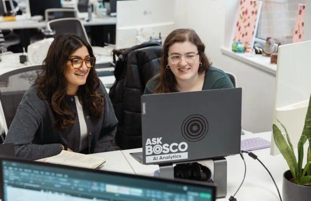 Two smiling women sit together at a desk, looking at a laptop displaying the Ask Bosco AI Analytics logo.
