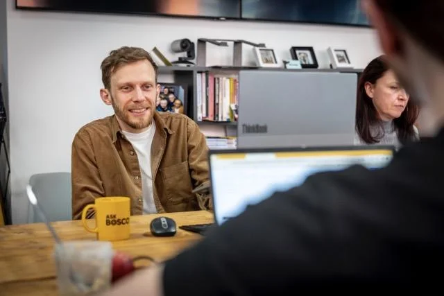 Man in brown shirt smiling during a meeting at a wooden table with a laptop and yellow mug.