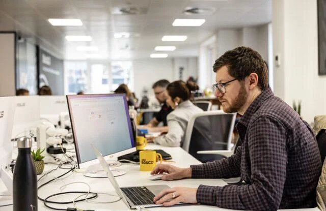 Man with glasses and beard working on a laptop at a desk in a modern open-plan office.