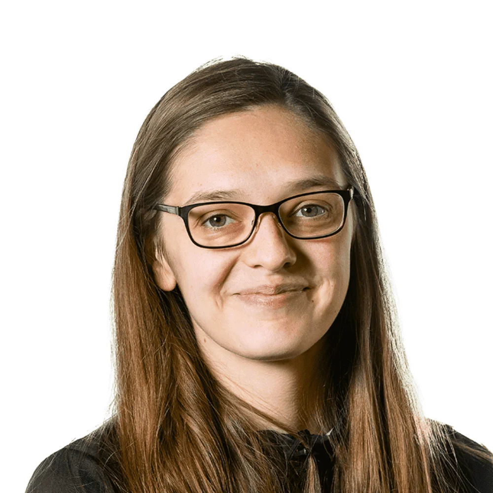 Professional headshot of a young woman with long brown hair, glasses, and a dark top, smiling softly.