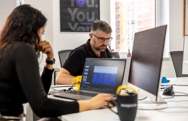 Man and woman working at desks with laptops and monitors in a modern office setting.