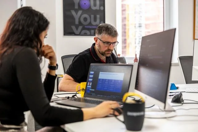 Man and woman working at desks with laptops and monitors in a modern office setting.