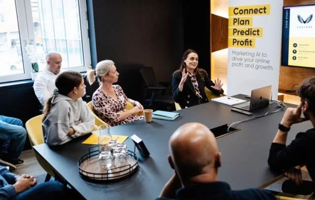 A woman gestures while speaking during a business meeting, with colleagues seated around a table beside a Marketing AI banner.