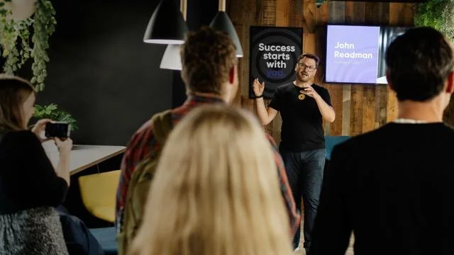 Speaker named John Readman gestures while presenting to an audience in an office, with a motivational poster behind him.