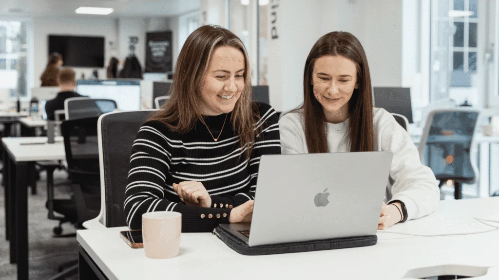 Two women smiling and collaborating at a laptop in a modern open-plan office.