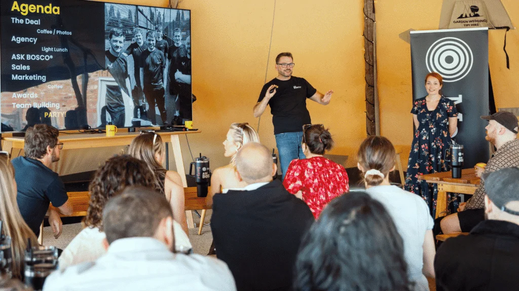 Man presenting company agenda on screen to seated audience in warm-lit venue with branded banner nearby.