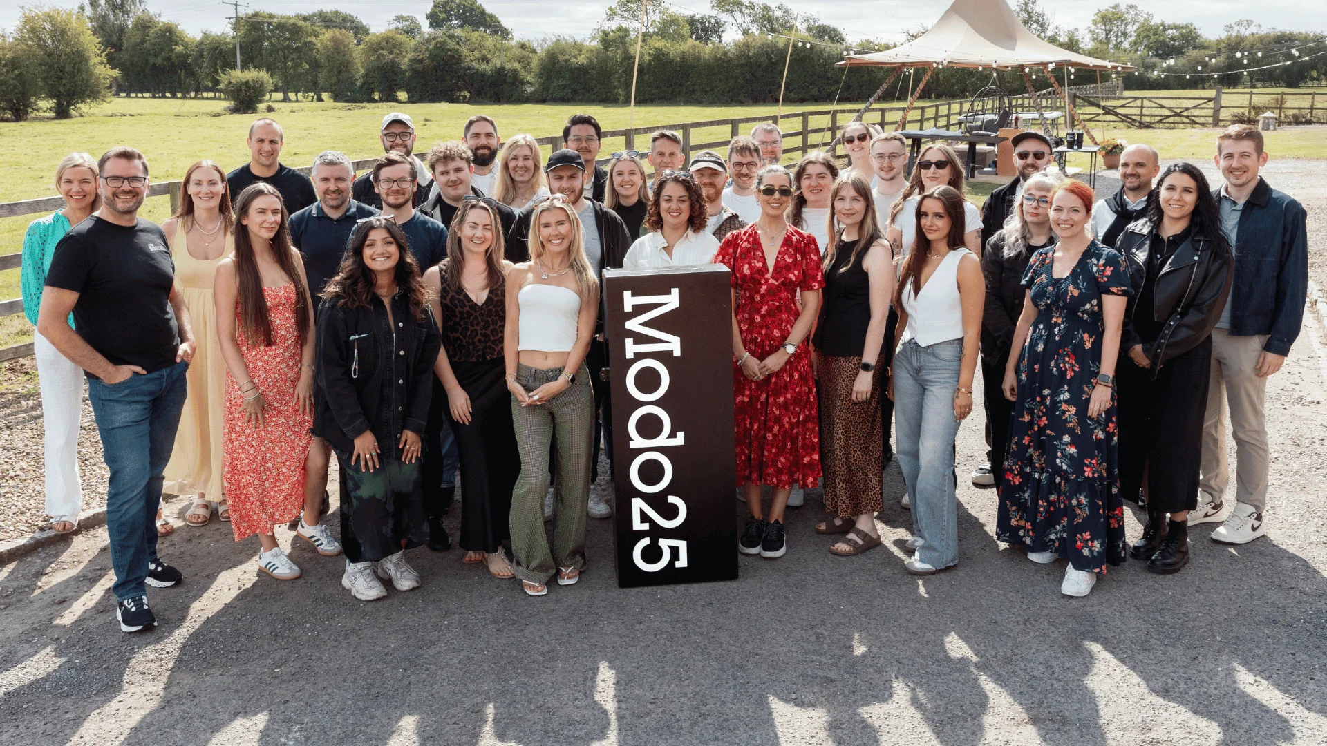 Large group of Modo25 employees posing together outdoors at a company event, gathered around the Modo25 branded sign.