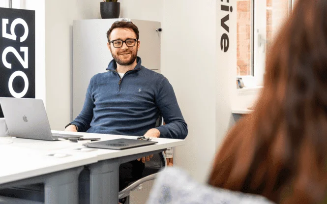 Man in glasses smiling during a meeting at an office desk with a laptop, engaging with a colleague.