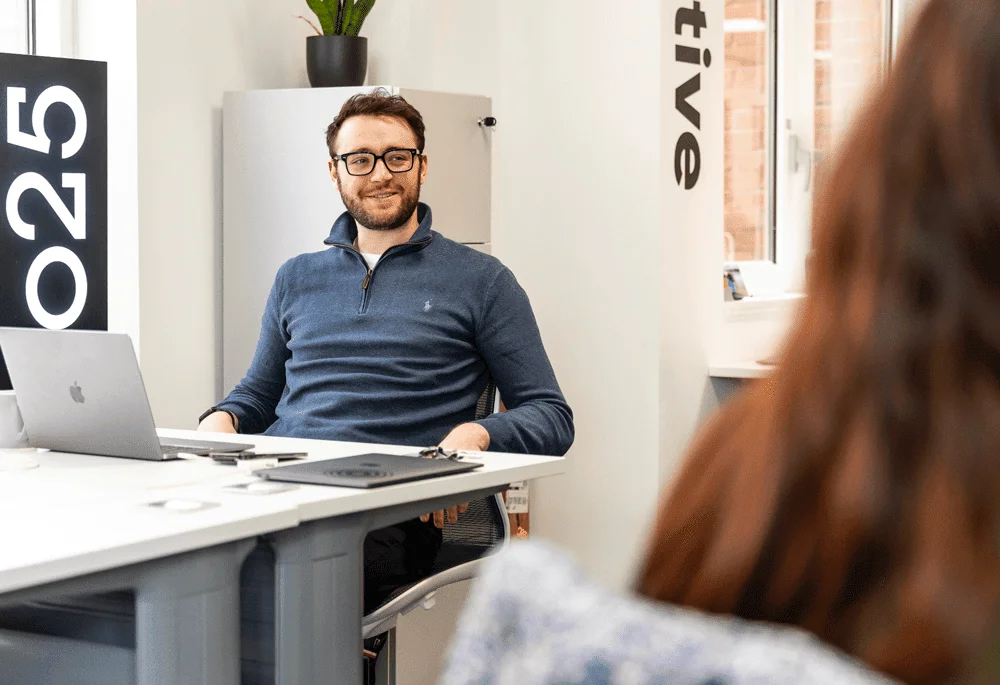 Man in glasses smiling during a meeting at an office desk with a laptop, engaging with a colleague.
