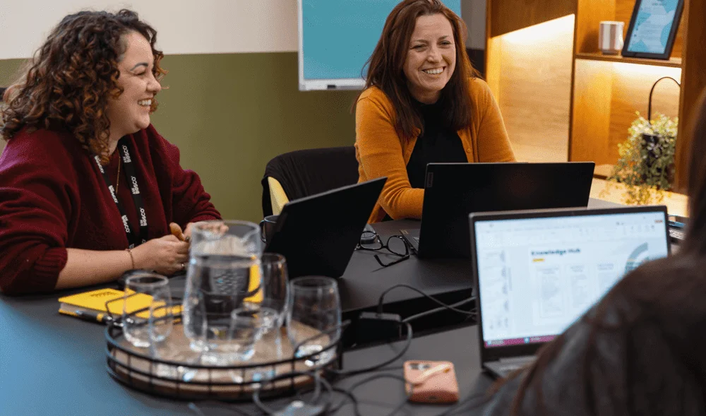 Two women smiling during an AB testing discussion at a laptop-filled meeting table.