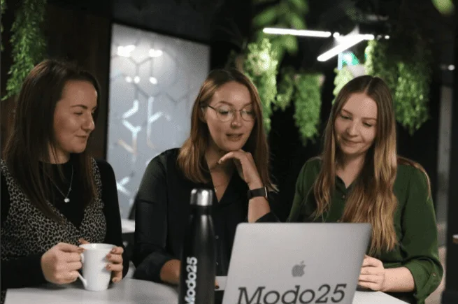Three women collaborating around a laptop with a Modo25 branded water bottle, in a plant-filled office setting.