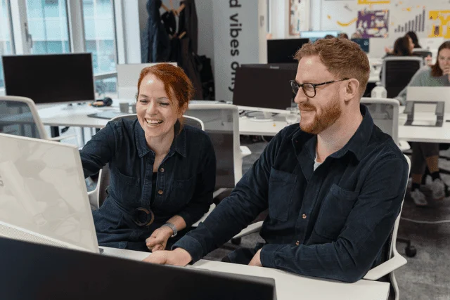 Two colleagues collaborating at a computer in a modern open-plan office, reflecting a supported in-housing work environment.