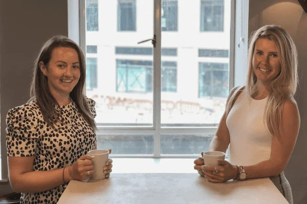 Modo 25 team members Kylie and Rebekah smiling and holding coffee mugs at a table by a window
