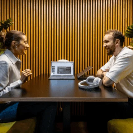 Two colleagues smiling in conversation at a table with a laptop and headphones, set against a wooden slat wall.