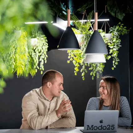 Two colleagues chat and smile while working together at a laptop under hanging plants and pendant lights.