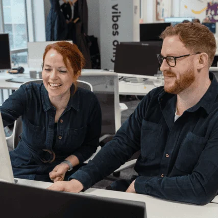 Two colleagues smiling and collaborating at a desk in a modern office workspace.