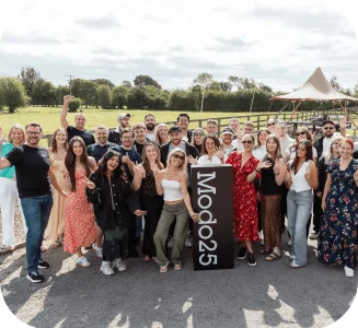 Large group of Modo25 employees smiling and posing together outdoors beside a branded sign