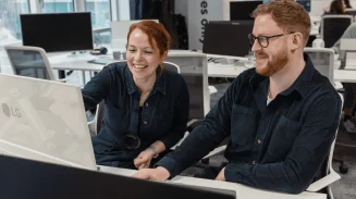 Two colleagues smiling while looking at a computer screen together in a modern open-plan office.