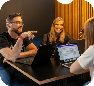Three colleagues smiling and discussing work around laptops during a casual office meeting.
