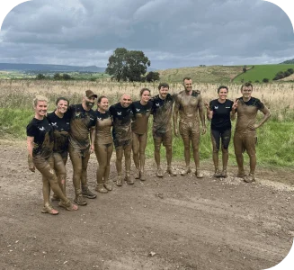 Group of nine mud-covered runners smiling together after completing an obstacle course race in a rural field.