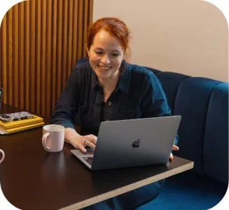 Smiling woman with red hair typing on a MacBook laptop at a wooden table with a coffee mug.